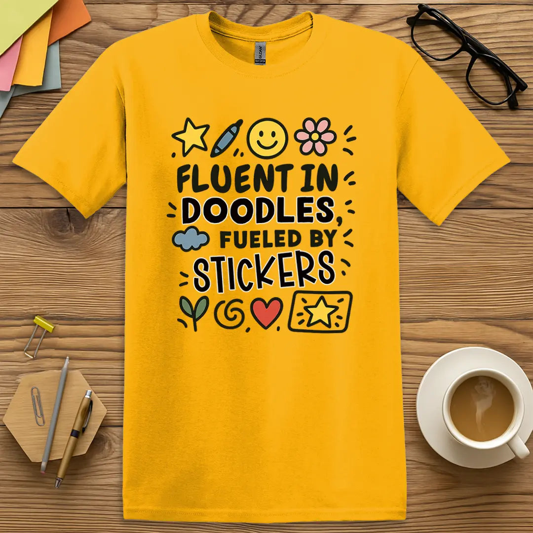 Gold Color T-shirt with "Fluent in Doodles, Fueled by Stickers" design featuring whimsical hand-drawn icons and classroom creativity vibe.